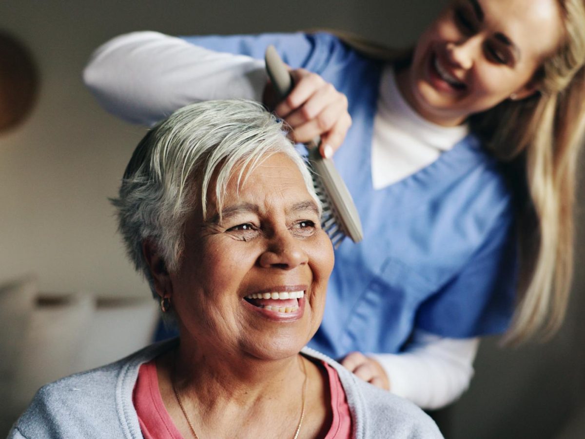 An older woman smiling as a carer gently combs her hair after washing, showing calm and dignified personal care.